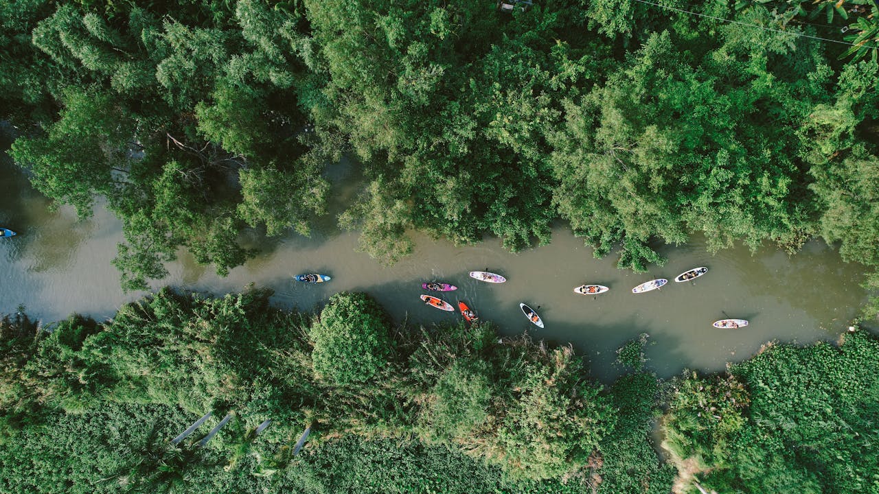 who-we-are A drone captures kayakers paddling through a serene river surrounded by dense green forest.
